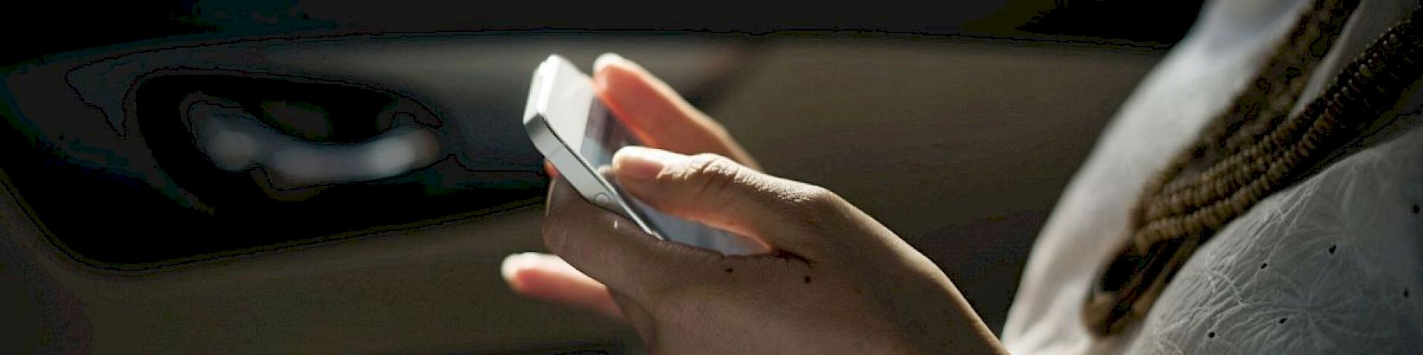 A person is holding and using a smartphone, possibly texting or browsing, inside a dimly lit car.