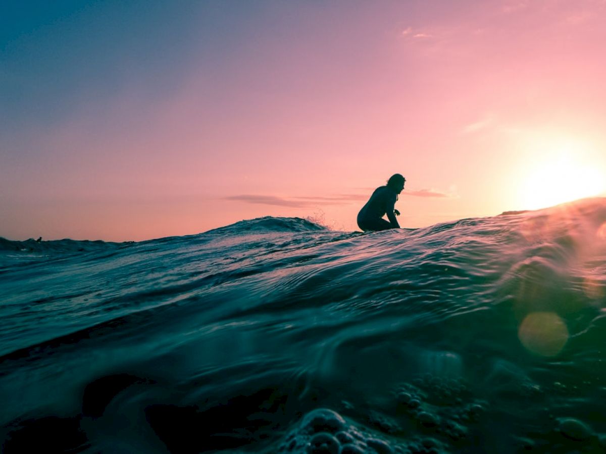 A silhouette of a person surfing on the ocean waves at sunset, with vibrant colors in the sky and a calm sea in the foreground.