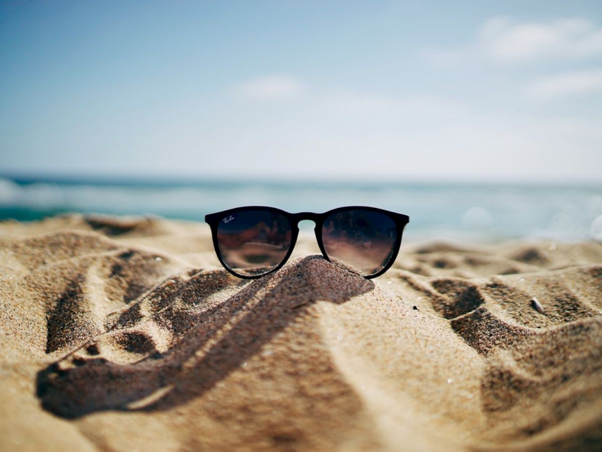 A pair of sunglasses on a sandy beach with the ocean in the background under a blue sky.