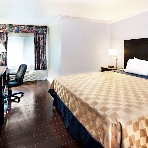 A neatly arranged hotel room with a large bed, desk with chair, and TV. There's a window with a curtain and a lamp on the side table.