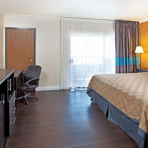 The image shows a hotel room with a bed, TV, desk, chair, lamp, and a window with curtains. The room has wood flooring and neutral decor.