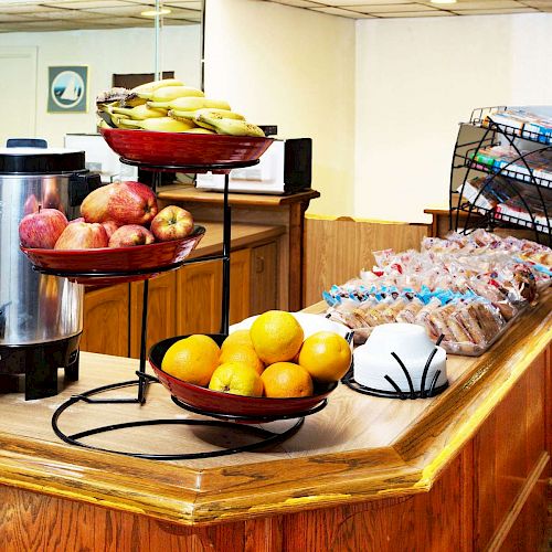 The image shows a breakfast buffet counter with fruits, wrapped pastries, a coffee dispenser, and an assortment of packaged snacks.