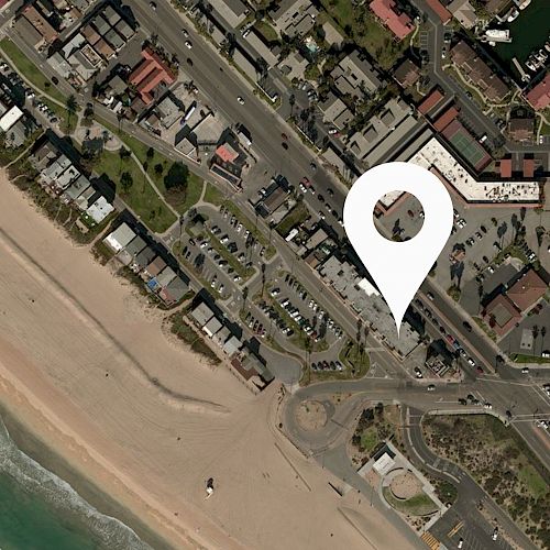 An aerial view of a beachside town with a location marker indicating a specific spot near the coast, showing streets, buildings, and a parking lot.