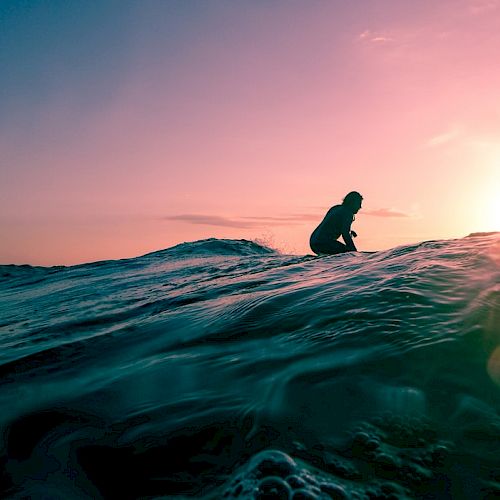 A silhouette of a person surfing on a wave with a vibrant sunset in the background, casting a beautiful mix of pink and blue hues on the water.