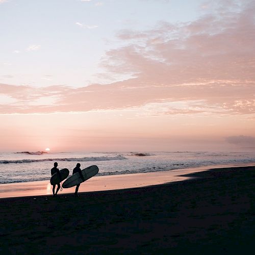 Two people carrying surfboards walk along a beach at sunset, with the ocean in the background and the sky filled with soft pink and orange hues.