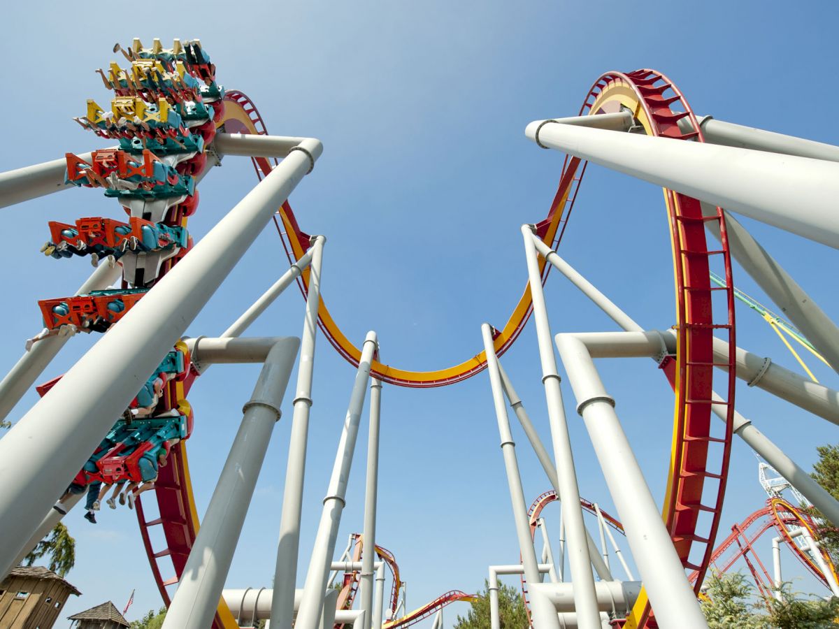 The image shows a roller coaster with loops and red and yellow tracks against a clear blue sky, with people riding the coaster.