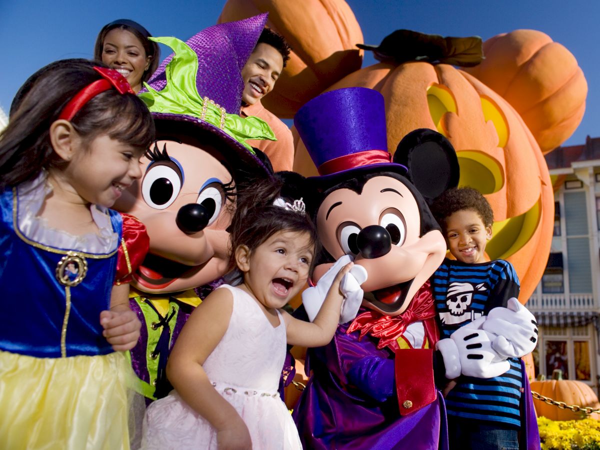 A group of children and adults are posing with costumed Mickey and Minnie Mouse characters in front of a large pumpkin decoration.