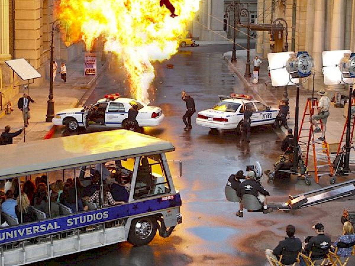 This image depicts a movie set with an explosion scene involving police cars, a film crew, equipment, and a Universal Studios tour bus.