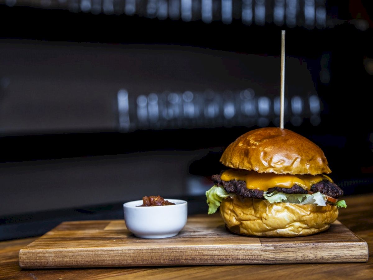 A juicy burger with melted cheese, lettuce, and a skewer on a board, alongside a small bowl of sauce, ready to be enjoyed.