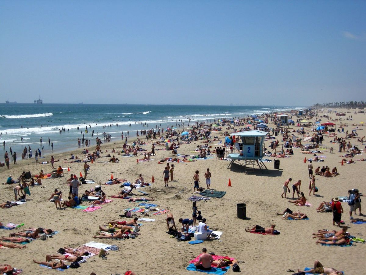 The image shows a crowded beach with many people sunbathing, swimming, and enjoying the sand near the ocean on a sunny day.