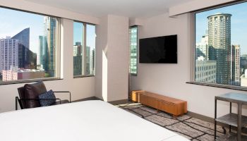 This image shows a modern apartment or hotel room with large windows offering a city view, a bed, a wall-mounted TV, and minimalist furniture.