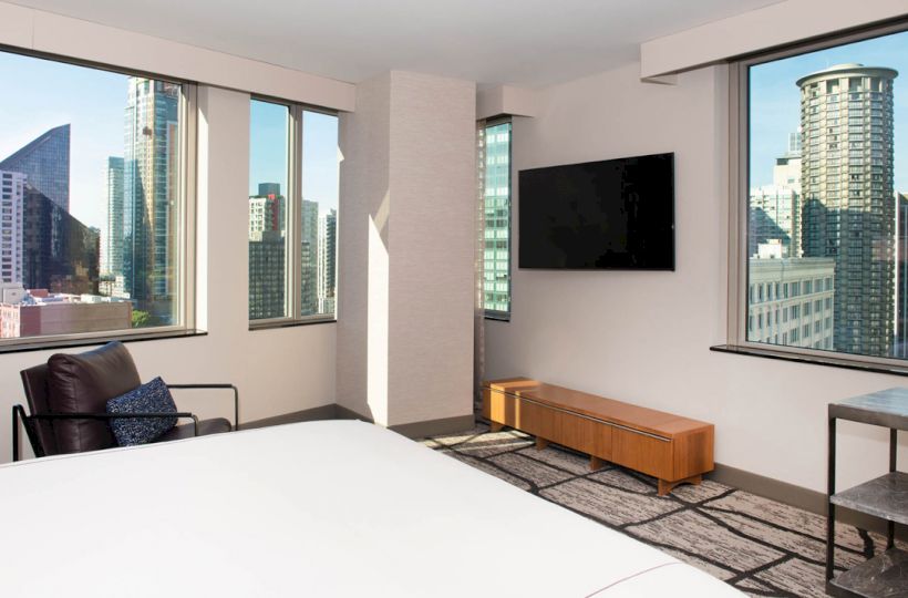 This image shows a modern apartment or hotel room with large windows offering a city view, a bed, a wall-mounted TV, and minimalist furniture.