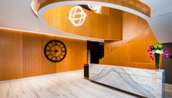 A modern lobby with a marble reception desk, wooden accent walls, a spiral staircase, a big wall clock, and a vase with flowers on the counter.