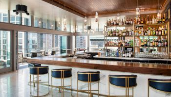A modern bar with high stools, a wooden counter, shelves stocked with various bottles, and large windows offering a city view ends the sentence.
