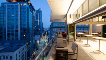 A modern outdoor terrace with seating overlooks a city street lined with tall buildings, captured during the evening.