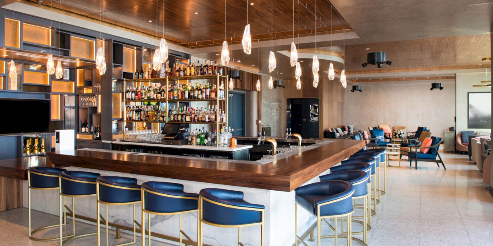 The image shows an upscale bar with a large counter, blue cushioned stools, hanging pendant lights, and a well-stocked selection of spirits.