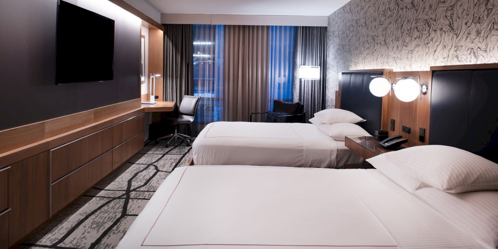 This image shows a modern hotel room with two beds, a large flat-screen TV, a work desk with a chair, and stylish decor.