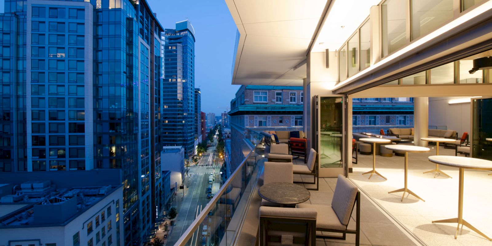 A modern cityscape with tall buildings is seen at dusk from a spacious balcony with tables, chairs, and an open indoor seating area.