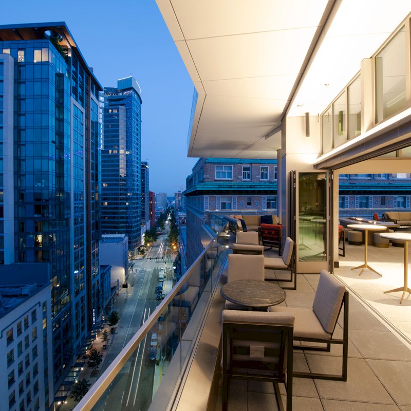 This image shows a modern high-rise cityscape at dusk, with a furnished balcony overlooking the street and adjacent buildings.
