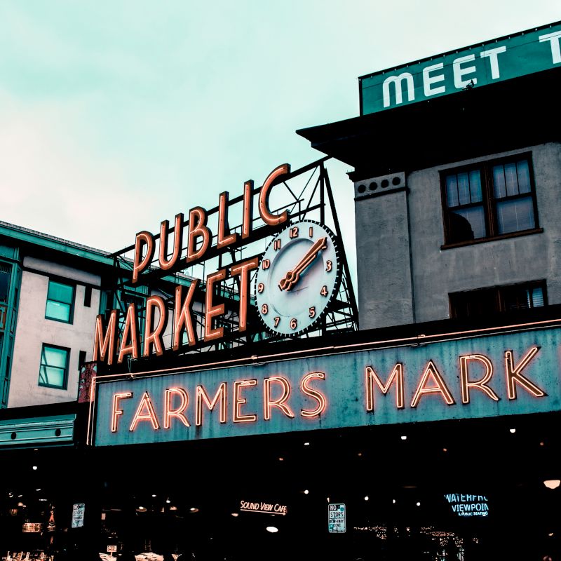 The image shows neon signs for "Public Market" and "Farmers Market" at a busy market area, with a clock and surrounding buildings in the background.