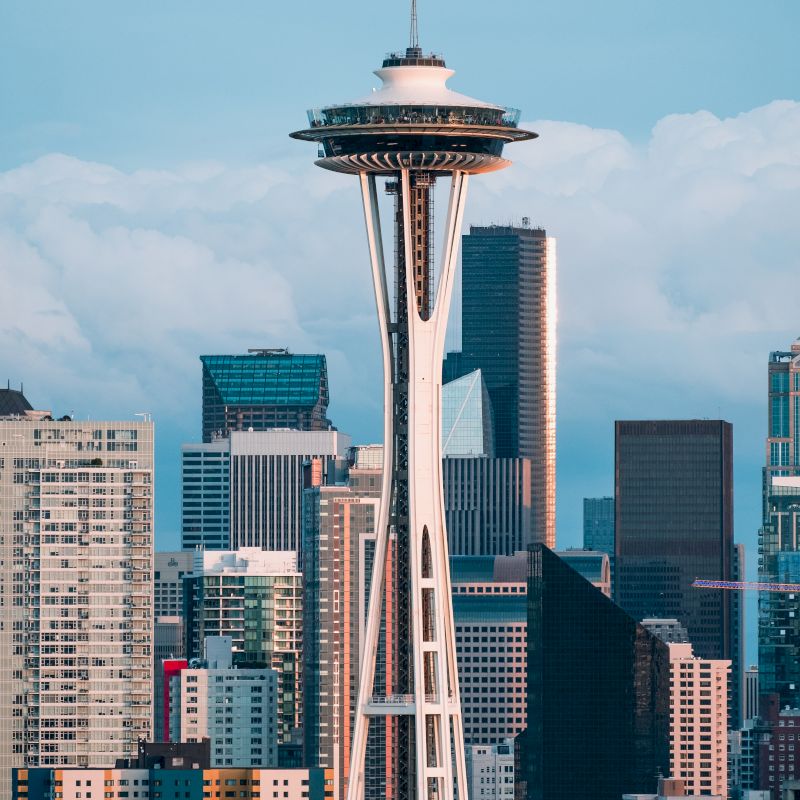A towering structure stands amid a cluster of skyscrapers, under a blue sky with clouds. The scene is a bustling urban landscape.