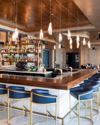 A modern bar with blue barstools, hanging lights, and a well-stocked shelf of bottles. The space also features seating areas and a flat-screen TV.