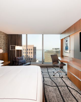 A modern hotel room with a large bed, a desk, a chair, a TV, and a window with a city and water view.