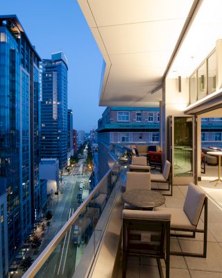 A modern cityscape with high-rise buildings is shown at dusk along with a sleek terrace that features outdoor seating and is partially illuminated.