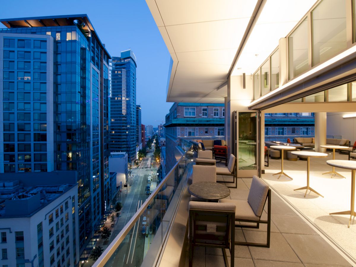 The image shows a modern city view during twilight from a high-rise balcony with outdoor seating areas, glass railings, and indoor lighting.