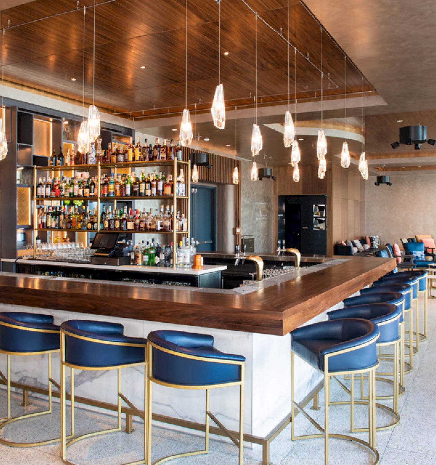 Modern bar with stylish lighting, well-stocked shelves, and blue barstools. Adjacent lounge area with seating and tables. Contemporary design.