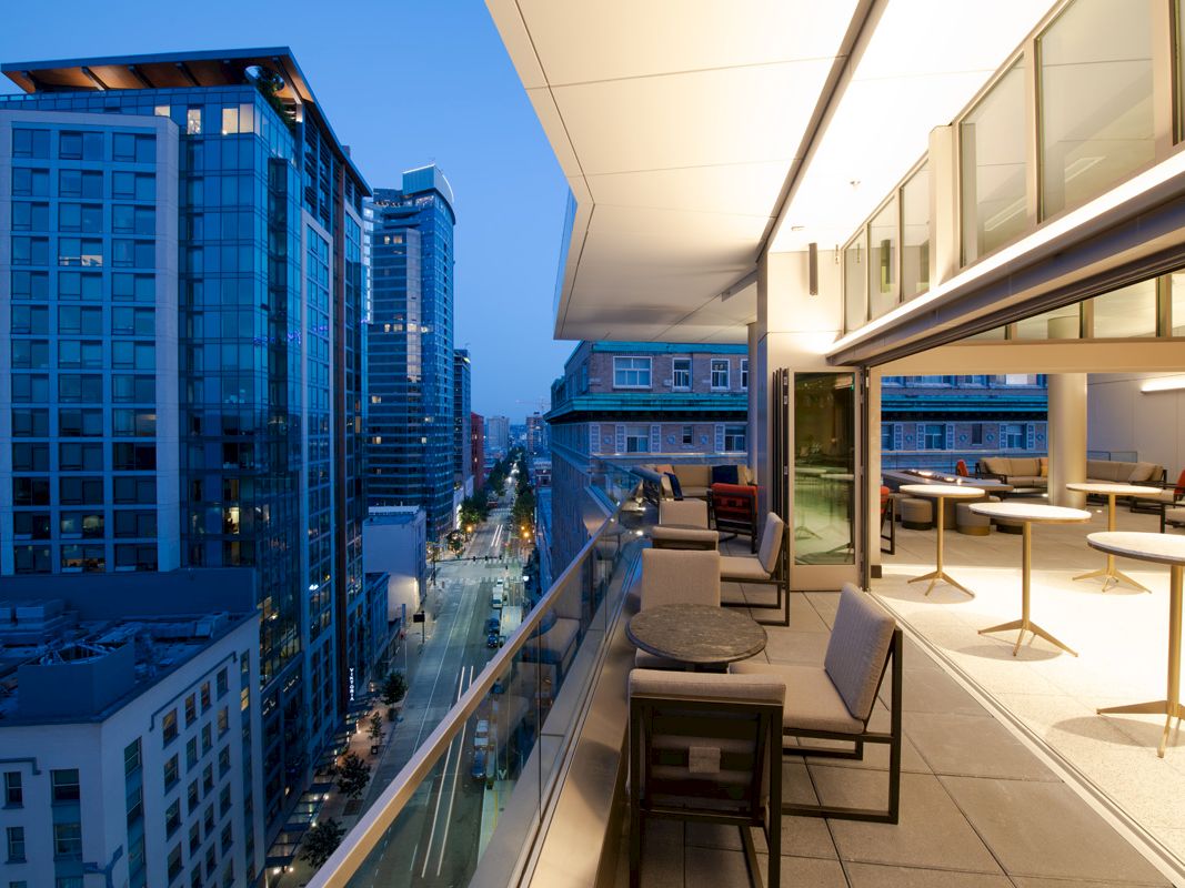 A modern balcony overlooks a cityscape with tall buildings. The balcony features seating areas and tables, lit by warm indoor lights as evening falls.