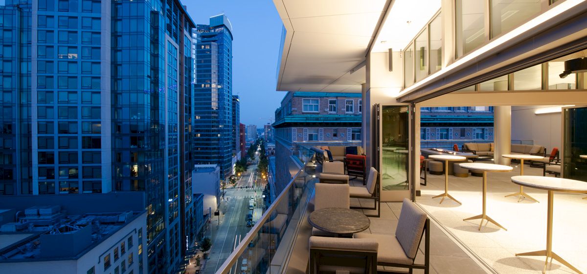 A modern balcony overlooks a cityscape with tall buildings. The balcony features seating areas and tables, lit by warm indoor lights as evening falls.