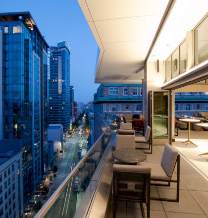The image shows a modern balcony with seating and tables overlooking a cityscape at dusk, featuring lit high-rise buildings and a street view.