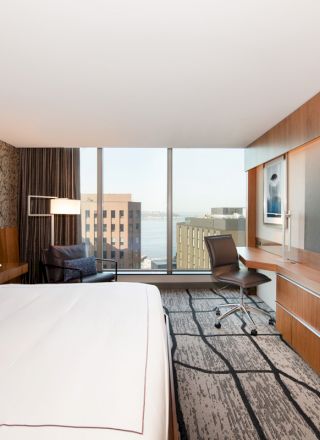 A modern hotel room with a large bed, a wall-mounted TV, a desk, a chair, and a window view of buildings and water outside the room.