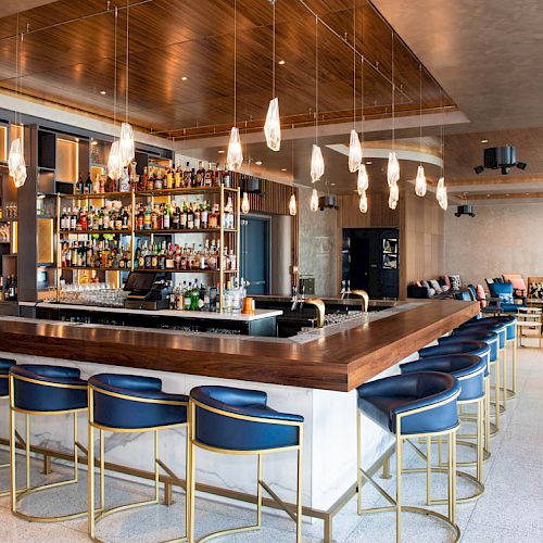 This image shows a modern bar with blue bar stools, a wooden bar counter, hanging pendant lights, and shelves stocked with bottles of alcohol.
