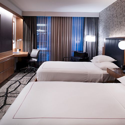 The image depicts a modern hotel room with two neatly made beds, a wall-mounted TV, a desk with a chair, and contemporary furnishings.