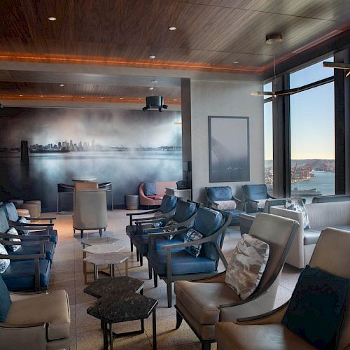 A modern lounge with comfortable seating, large windows showcasing a waterfront view, a mural on the wall, and plenty of natural light.