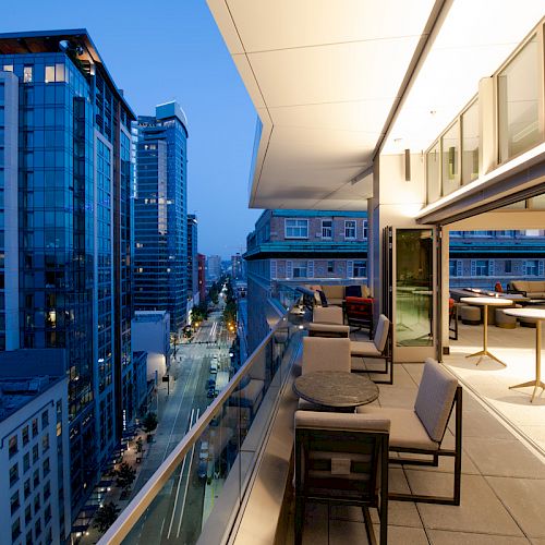A modern cityscape as seen from a balcony at dusk, featuring tall buildings, street lights, and a luxurious outdoor seating area.
