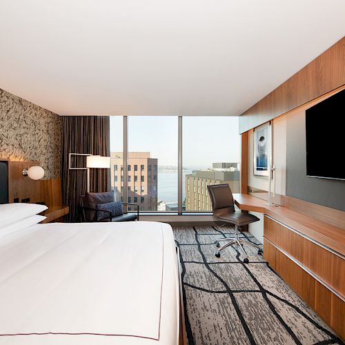 A modern hotel room with a large bed, a wall-mounted TV, a desk, chair, and a view of buildings and water through a large window.