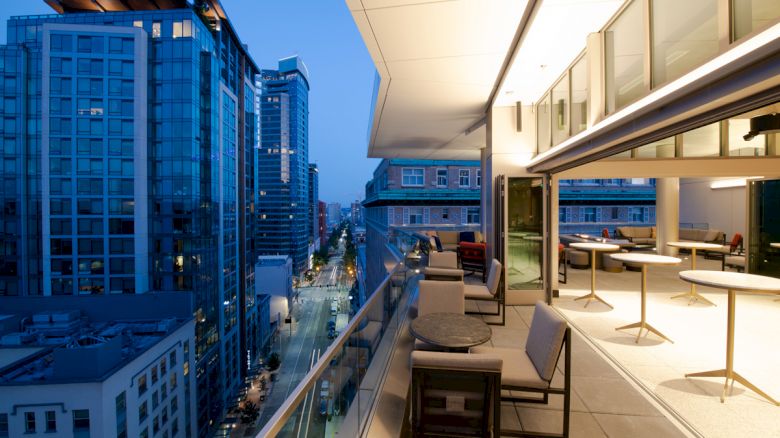 An evening cityscape from a modern balcony with outdoor seating and table arrangements, overlooking high-rise buildings and a lit-up street.