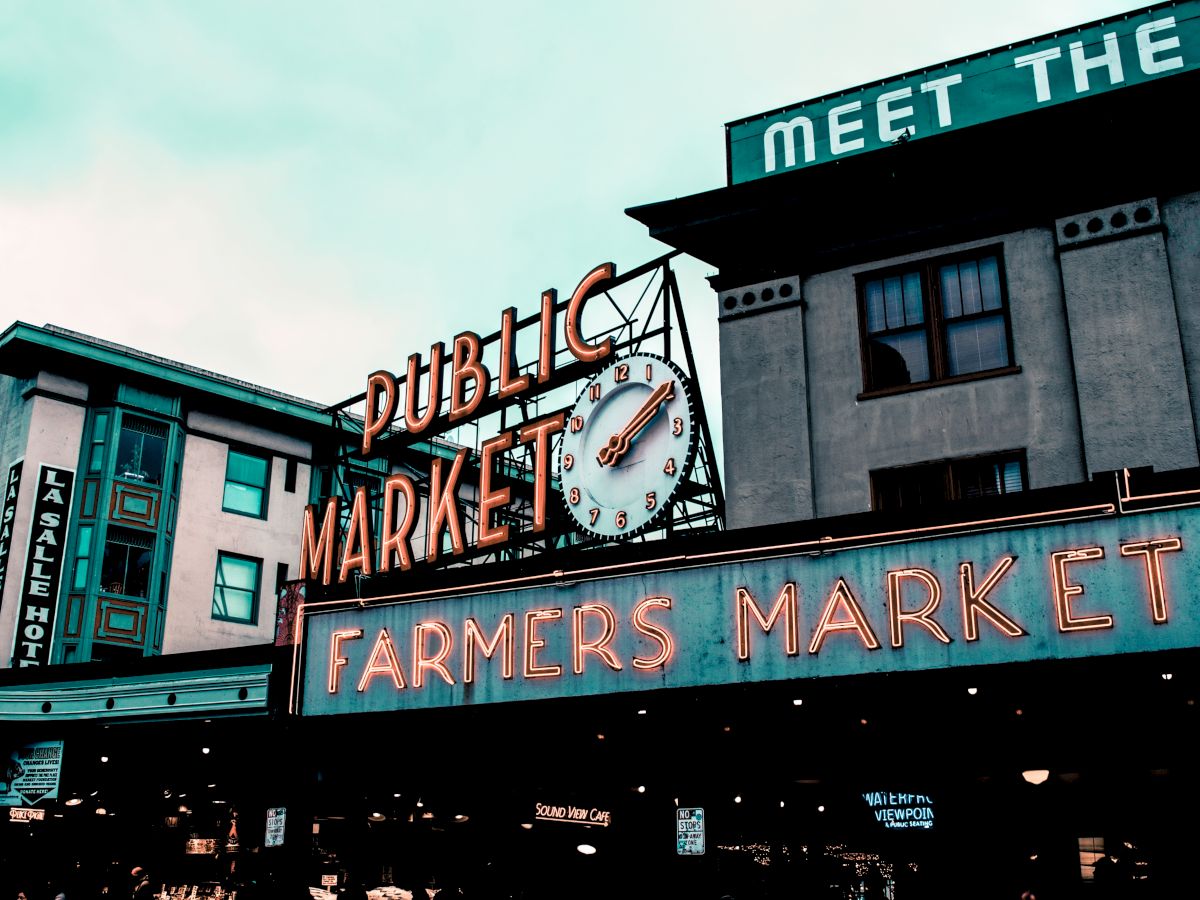Image of a public market with neon signs reading "PUBLIC MARKET" and "FARMERS MARKET" on a busy street, with buildings in the background.