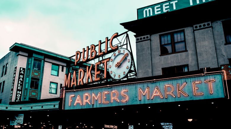 Image of a public market with neon signs reading "PUBLIC MARKET" and "FARMERS MARKET" on a busy street, with buildings in the background.