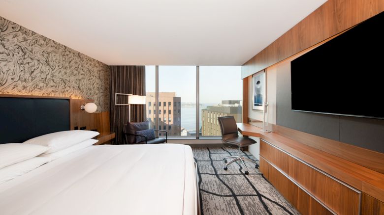 A modern hotel room with a large bed, a TV, a desk and chair, and a window offering a city view outside.