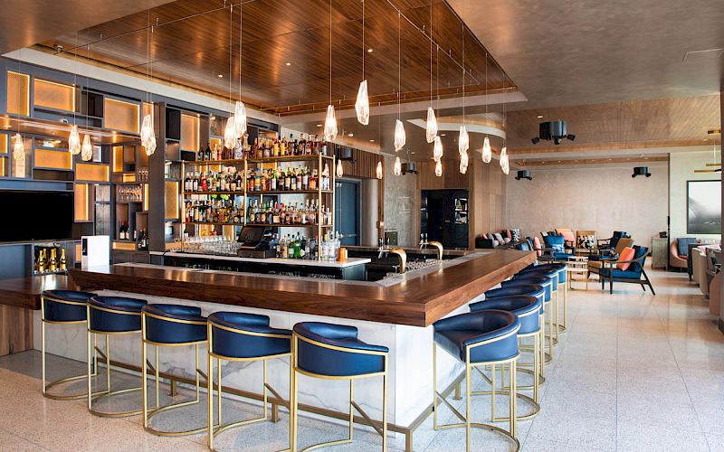 A modern bar with high stools, a well-stocked liquor shelf, hanging lights, and a stylish seating area in the background.