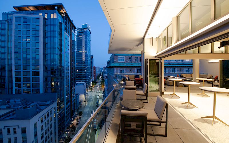 A modern outdoor balcony with seating and tables overlooks a cityscape with tall buildings at dusk. The view is both urban and serene.