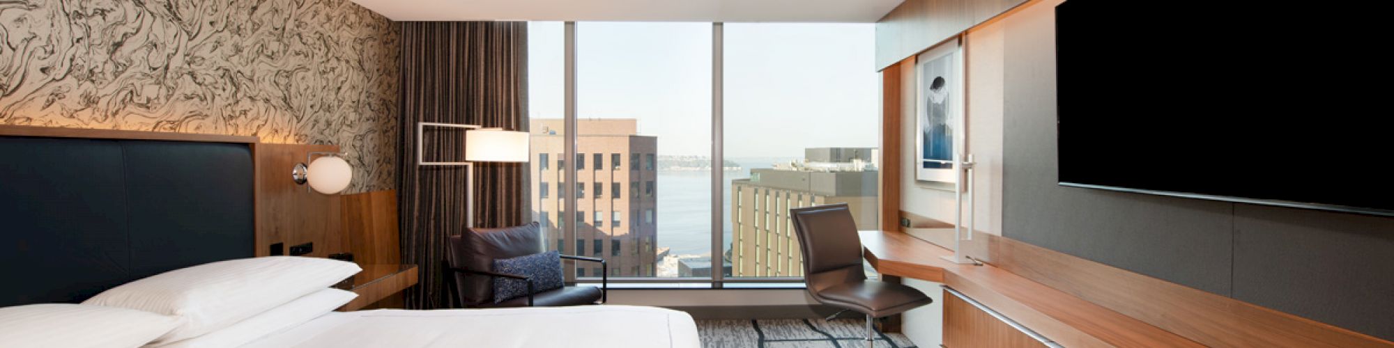 A modern hotel room featuring a large bed, desk, chair, TV, and a window with a cityscape view in the distance, over a patterned carpet.