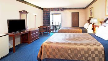 The image shows a hotel room with two beds, a TV on a dresser, a desk, a chair, and a lamp. There is a window with curtains and a door.