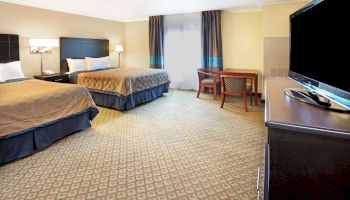 The image shows a hotel room with two double beds, a flat-screen TV, a small table with two chairs, and a window with curtains.