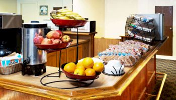 A breakfast buffet with fresh oranges, bananas, apples, pastries, and a beverage station with a coffee dispenser is shown in the image.