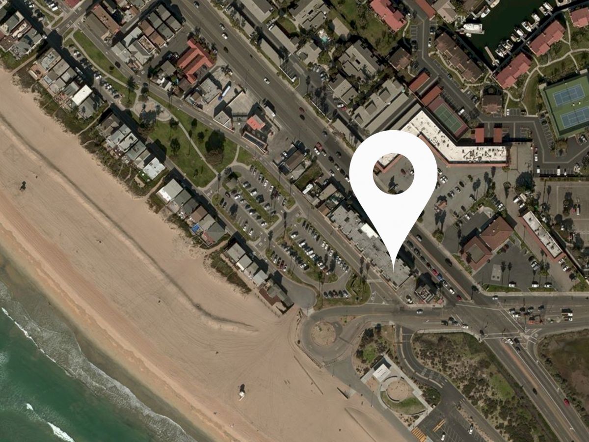 An aerial view of a coastal town with a beach, roads, buildings, and a large map pin marker indicating a specific location in the area.